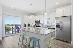 Una cocina moderna con armarios blancos, un refrigerador de acero inoxidable, una isla de granito con taburetes y una puerta corrediza de vidrio al exterior.
