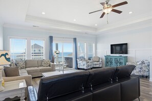 A spacious living room with a large black sofa, a television, and a ceiling fan. -    Luxurious Oceanside w/pool, elevator, 6 BR, steps from the sand and surf  (North Topsail Beach)