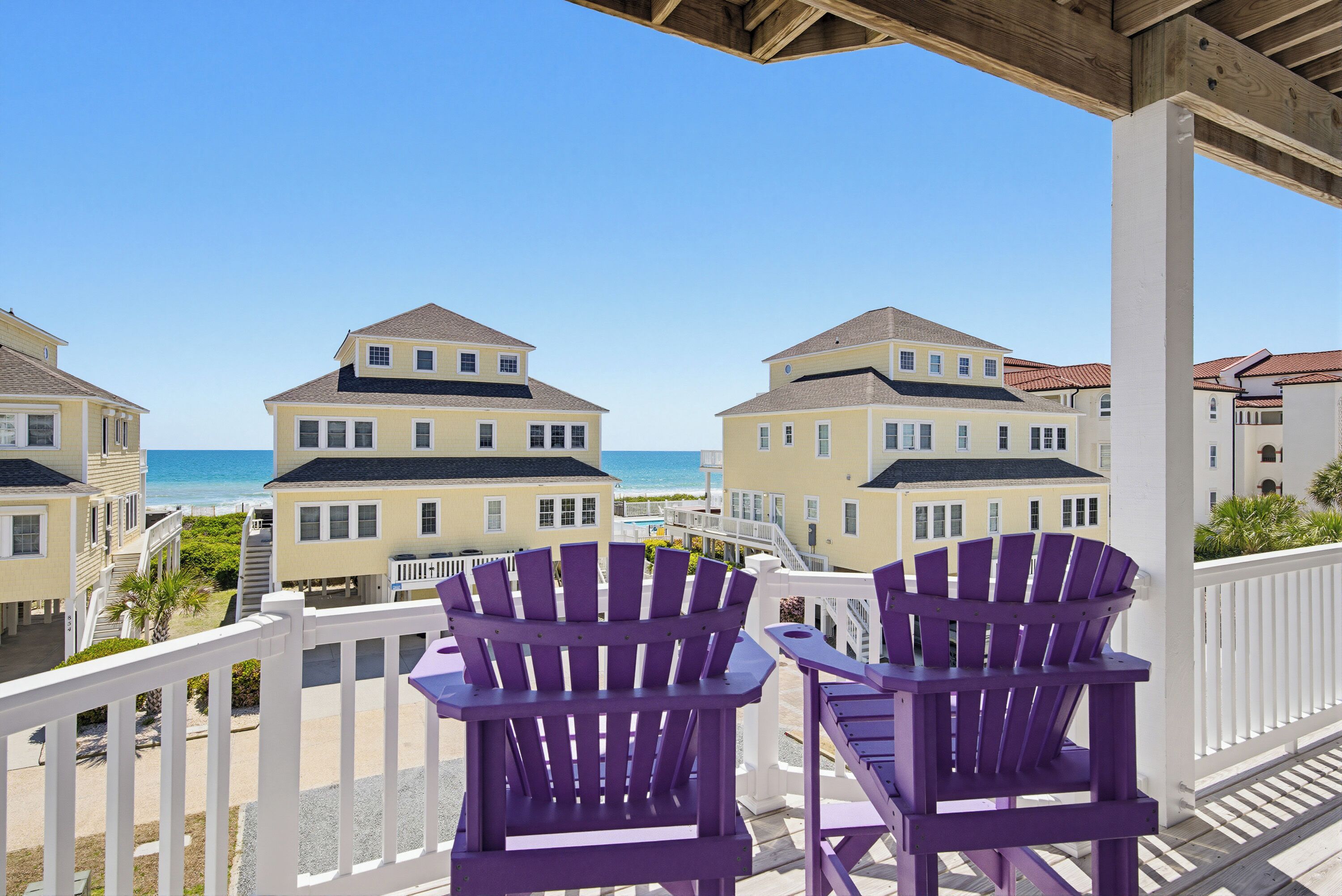 A balcony with two purple Adirondack chairs overlooking a beach and a row of yellow buildings.