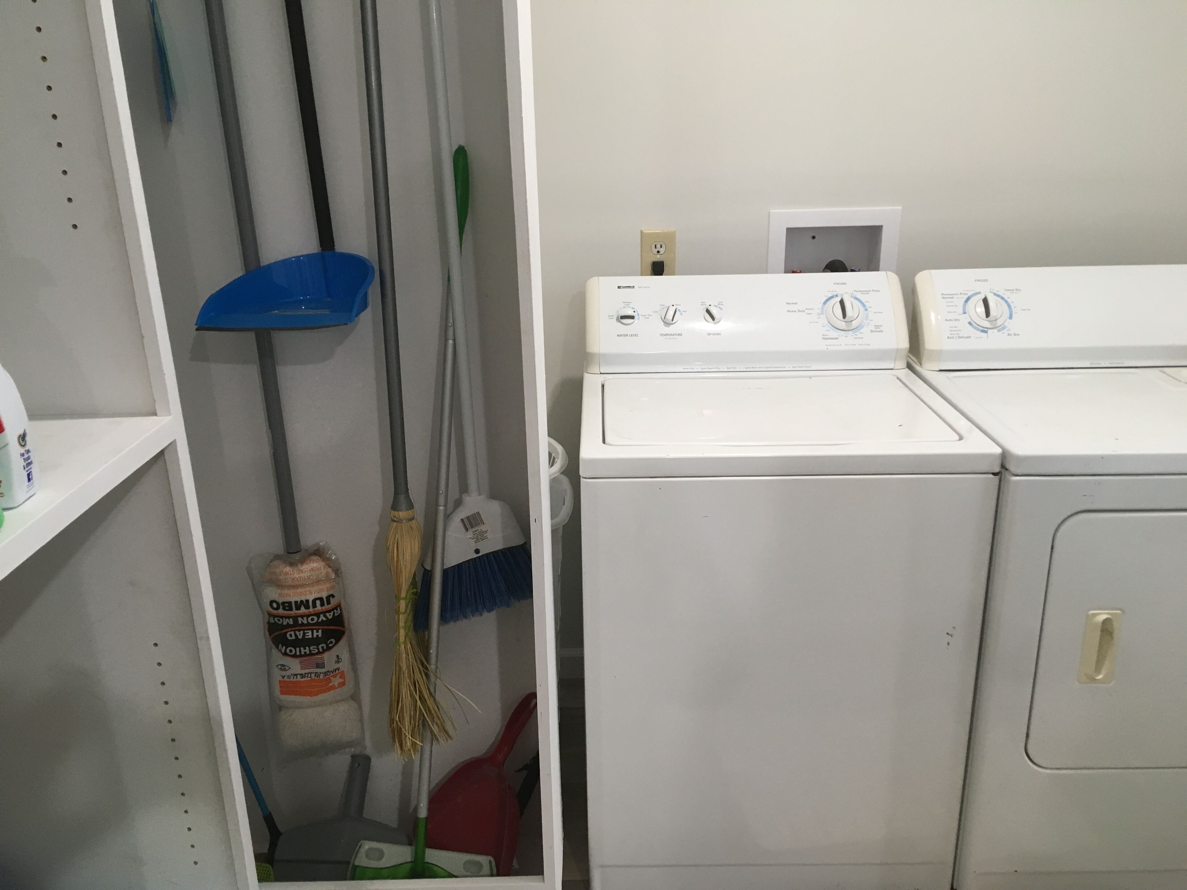 A laundry room with two stacked washing machines, a broom, a dustpan, and a container of laundry detergent.