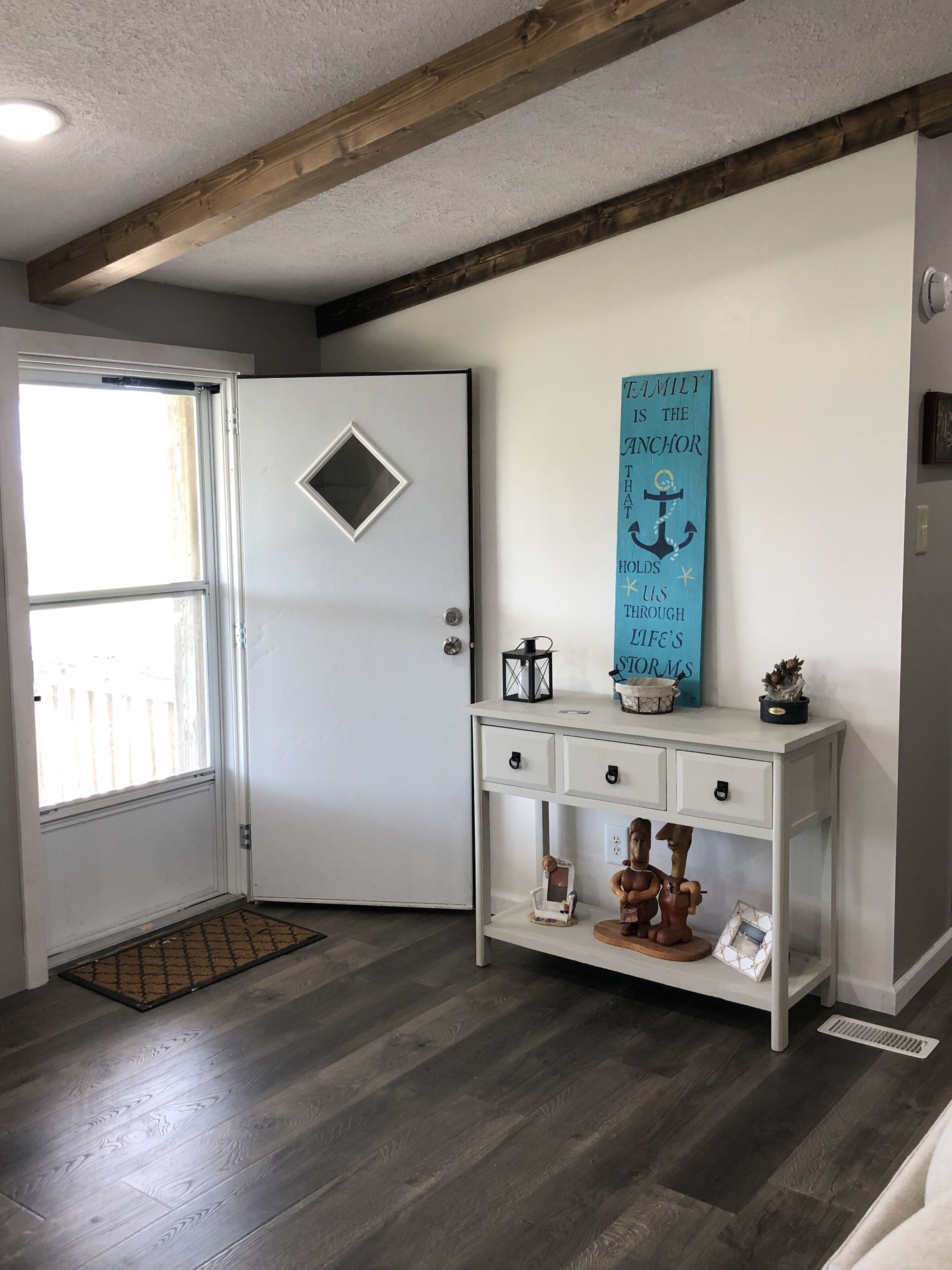 Une pièce avec une porte blanche, un plafond en bois, une console blanche avec des tiroirs, un panneau décoratif en forme d’ancre et une fenêtre donnant sur un paysage agréable.