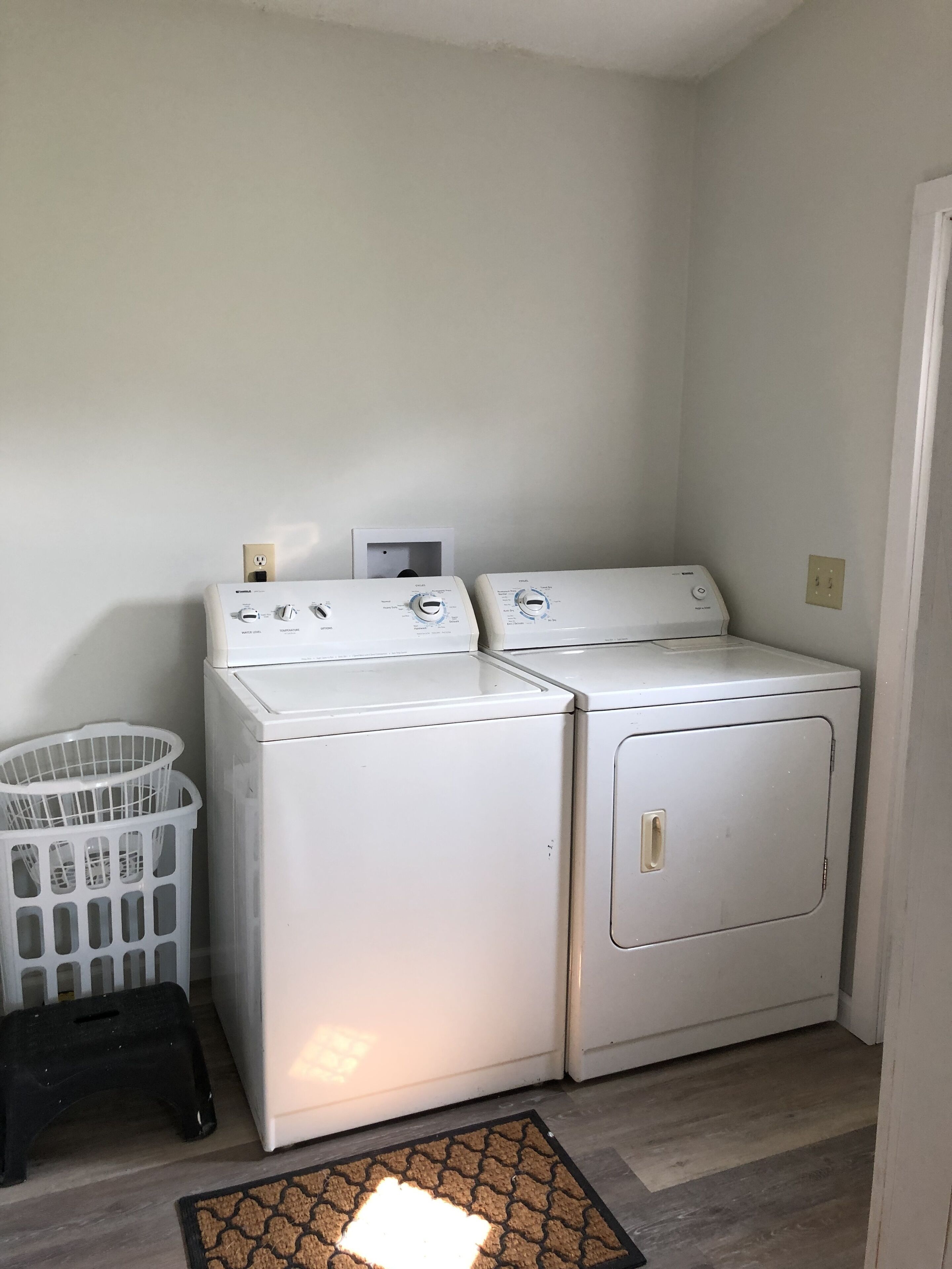 A laundry room with two white front-loading washing machines, a white plastic laundry basket, and a black stool.