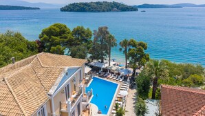 A pool area with lounge chairs and umbrellas, surrounded by trees and buildings with tiled roofs. - Hotel Orion (Lefkada)