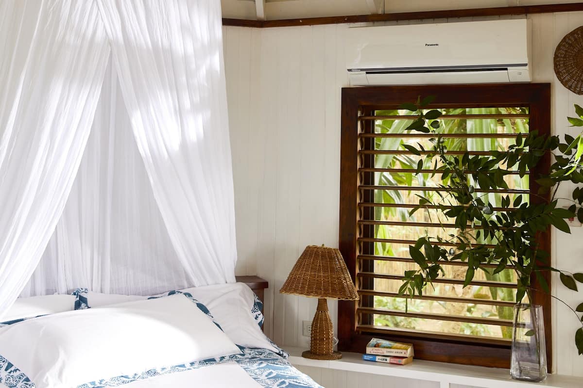 Une chambre à coucher avec un lit à baldaquin, une fenêtre donnant sur un paysage verdoyant, une lampe de chevet et un climatiseur fixé au mur.