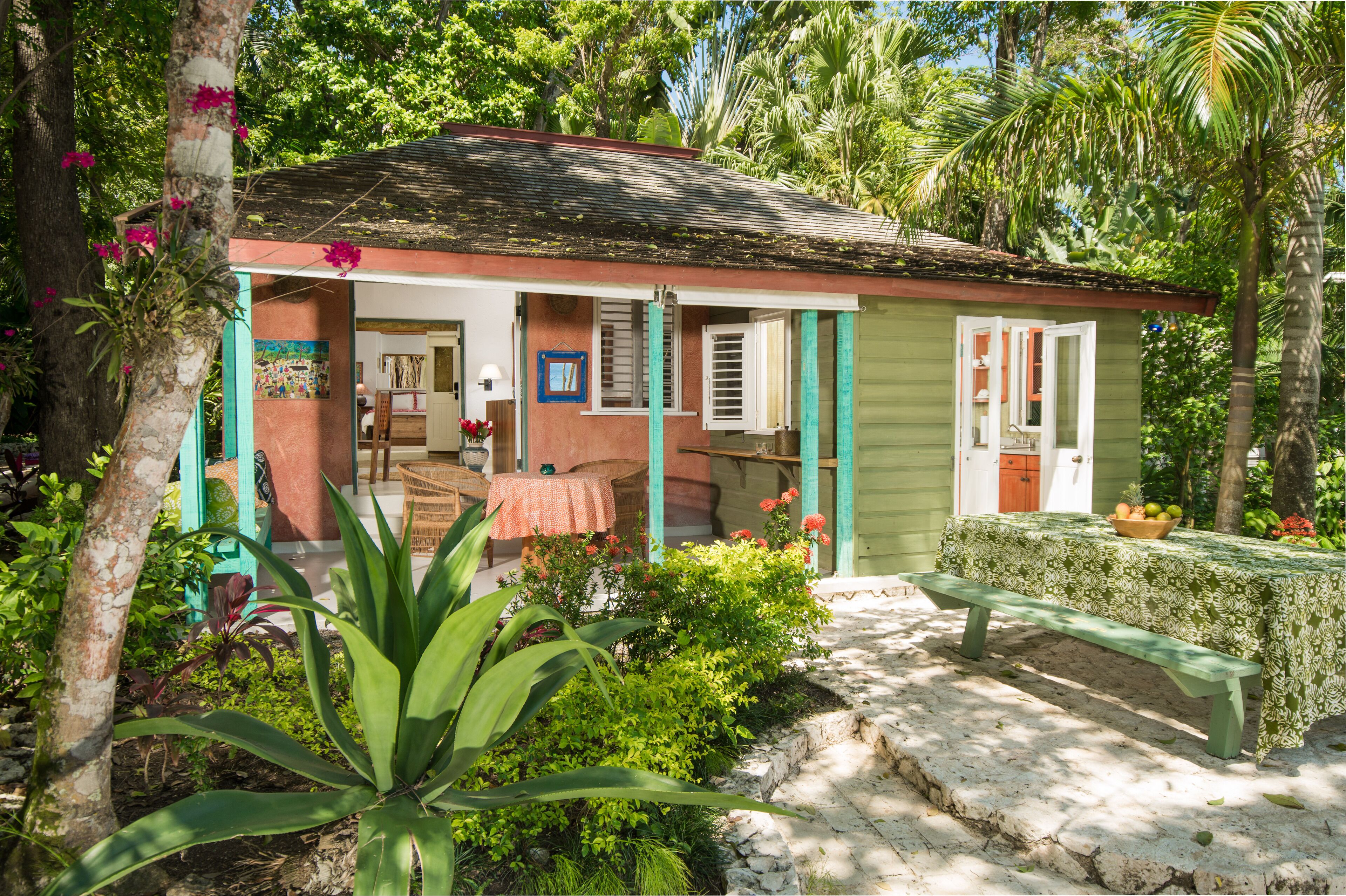 A small, single-story house with a porch, surrounded by lush greenery and a stone pathway.