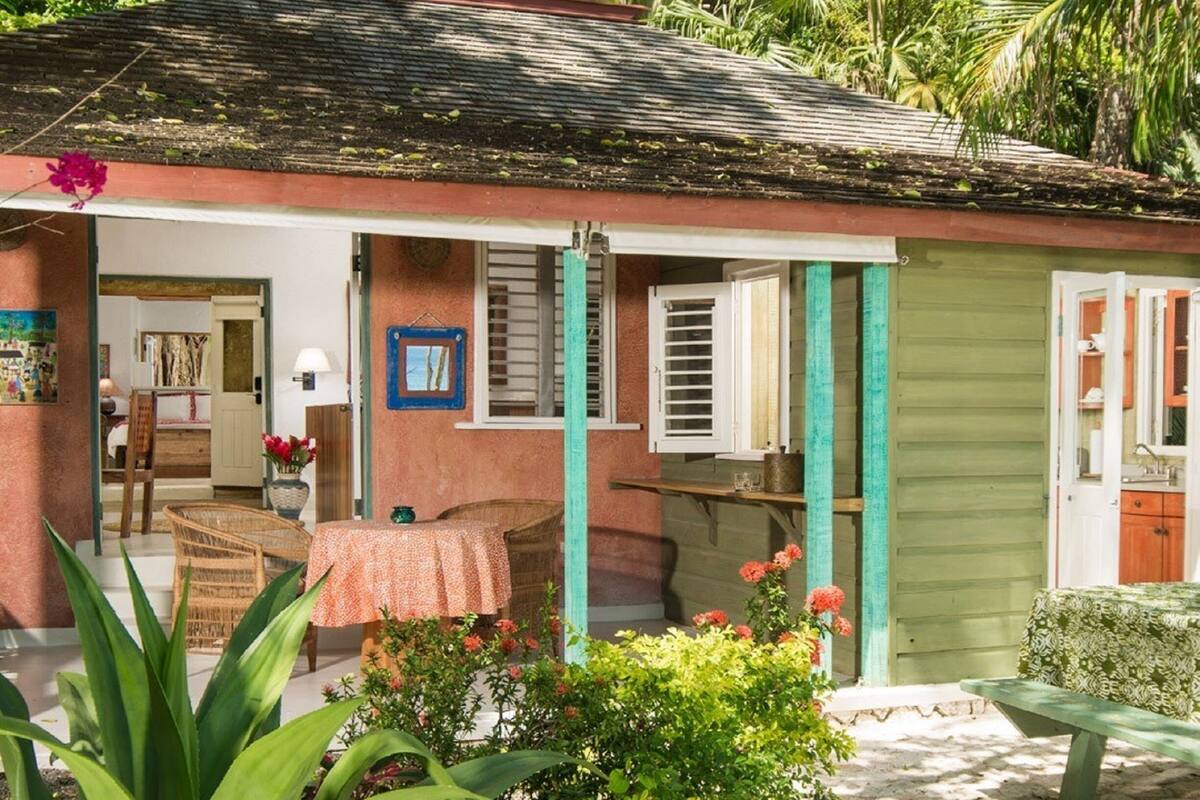 Une petite maison de plain-pied, avec un toit recouvert de tuiles, une véranda dotée d’une table et de chaises, et une porte verte.