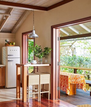 A compact kitchen with a white refrigerator, a hanging pendant light, wicker chairs, and a view of a garden through an open door.