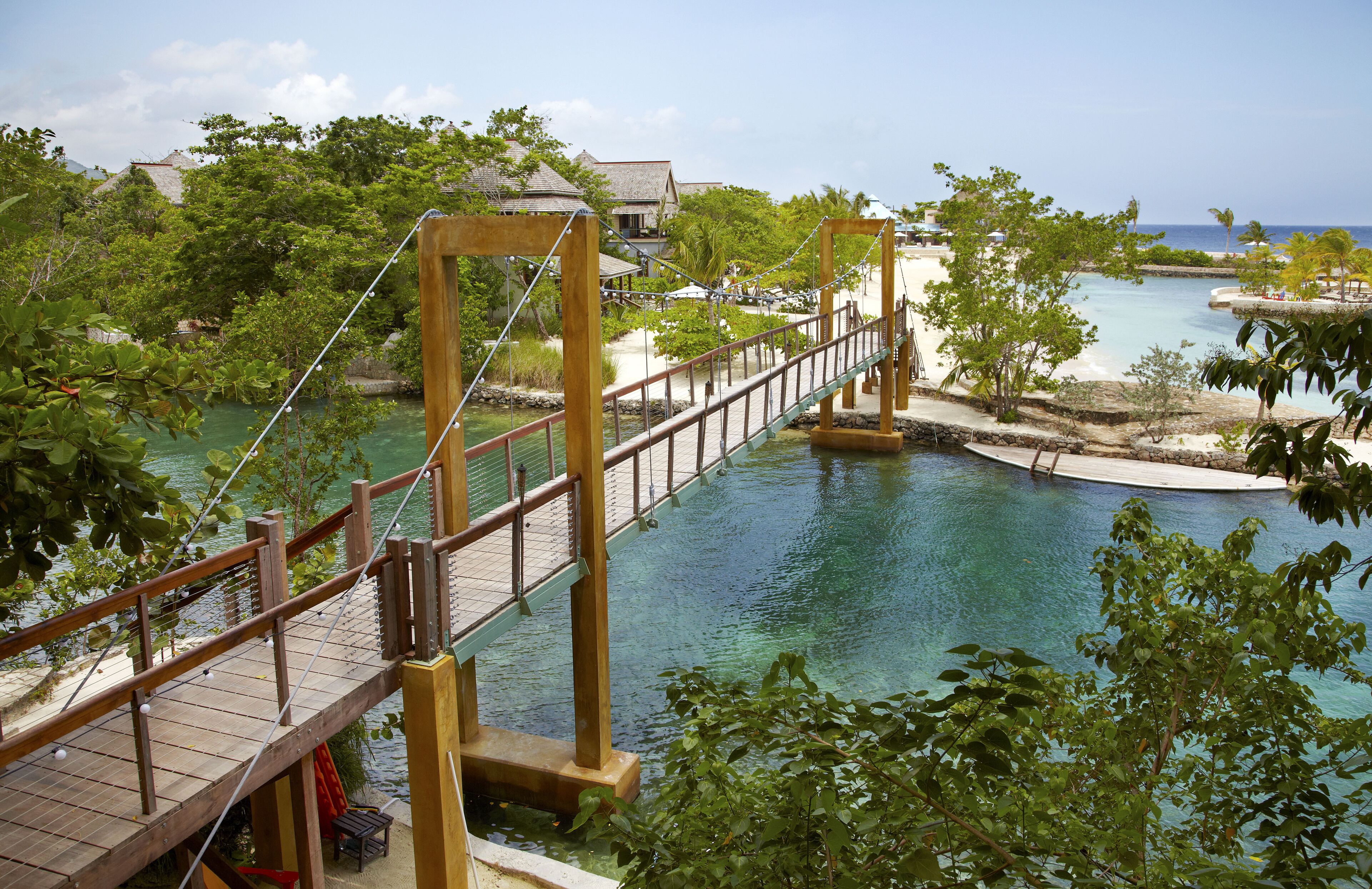 A wooden suspension bridge over a clear blue water body, surrounded by lush greenery and a few buildings in the background.
