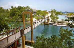 A wooden suspension bridge over a clear blue water body, surrounded by lush greenery and a few buildings in the background.