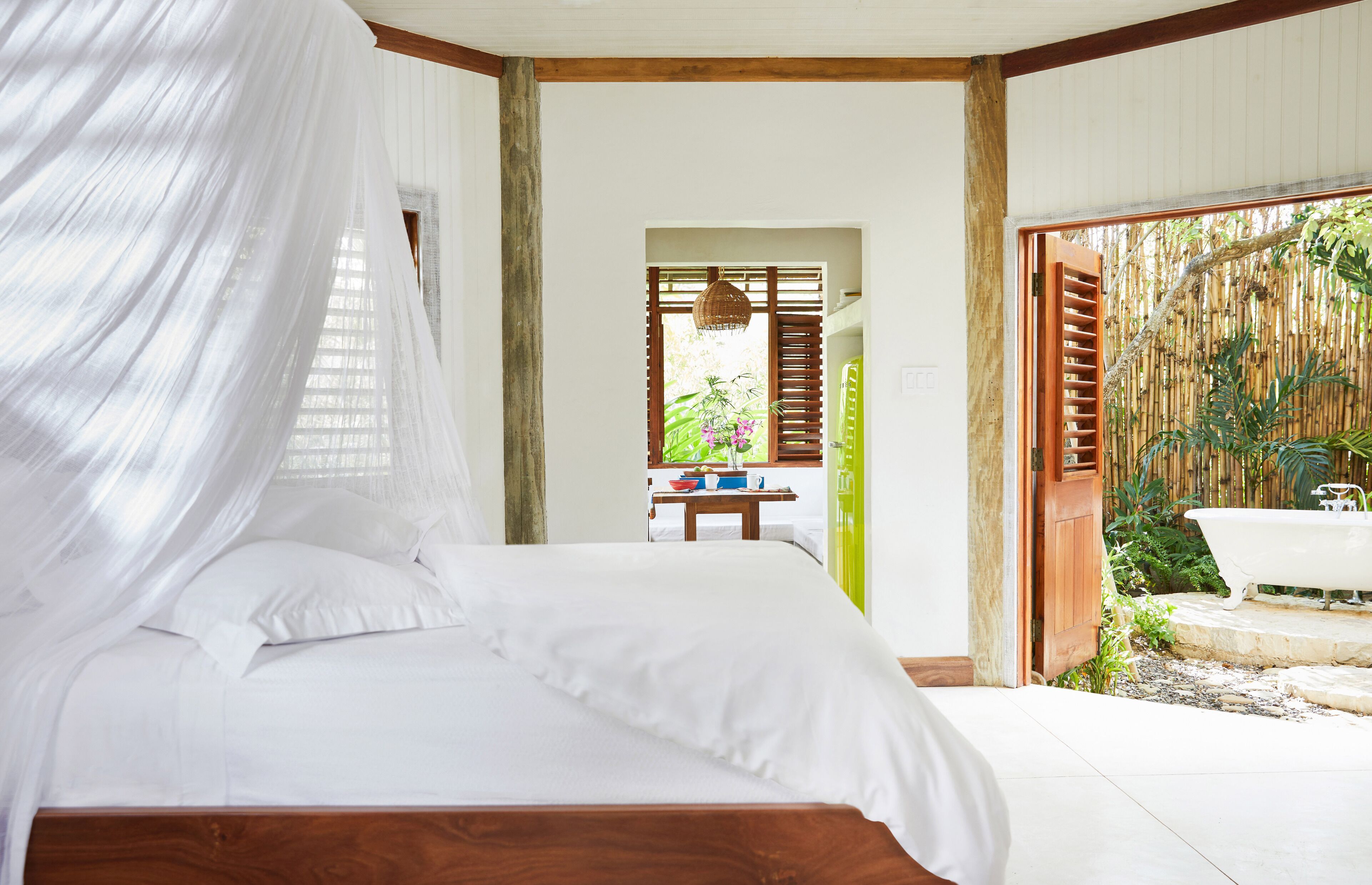 Ein Schlafzimmer mit einem Bett, einem Holz-Kopfteil, einem Fenster mit weißem Vorhang und Blick auf einen Bambuszaun sowie eine Badewanne im Freien.