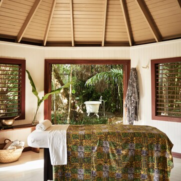 A room with a wooden ceiling, a bed with a patterned bedspread, a hanging bathrobe, and a view of a natural landscape through a window.