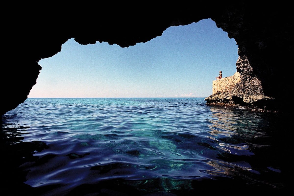 Vista a través de una cueva hacia un mar azul cristalino y un promontorio rocoso con una persona de pie sobre él.