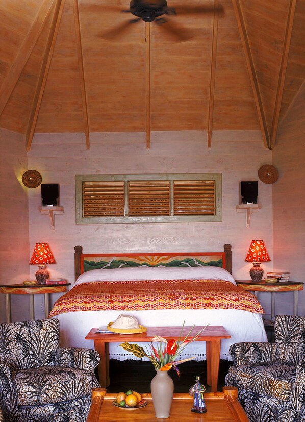 A cozy room with a wooden ceiling, a bed with a patterned bedspread, two lamps, a vase with flowers, and patterned chairs.