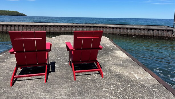 Zwei rote Adirondack-Stühle blicken auf einen Steg, der sich über ein Gewässer erstreckt, mit blauem Himmel darüber.