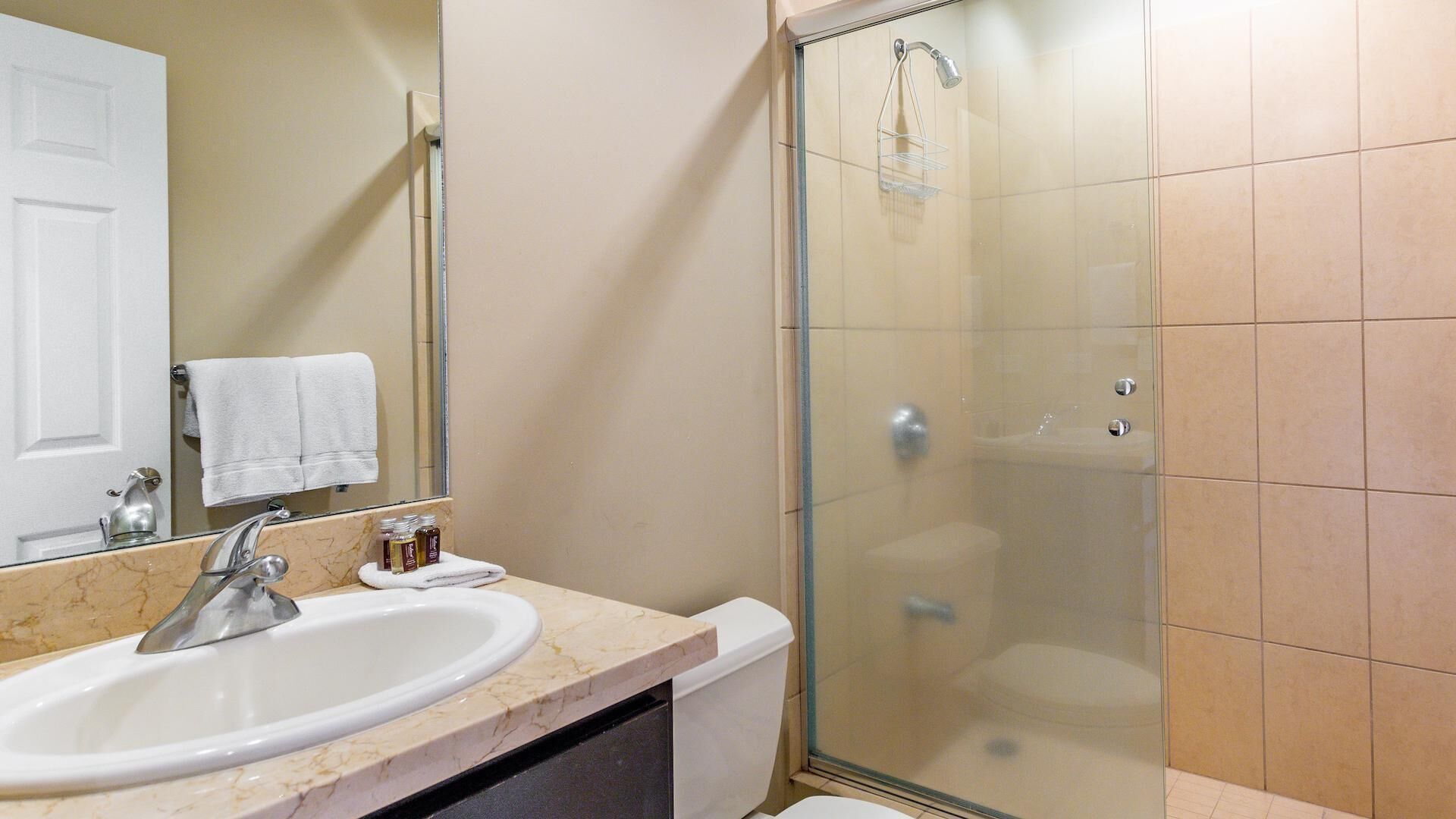 Een badkamer met een witte wasbak, een spiegel, een glazen douchecabine en beige tegels.
