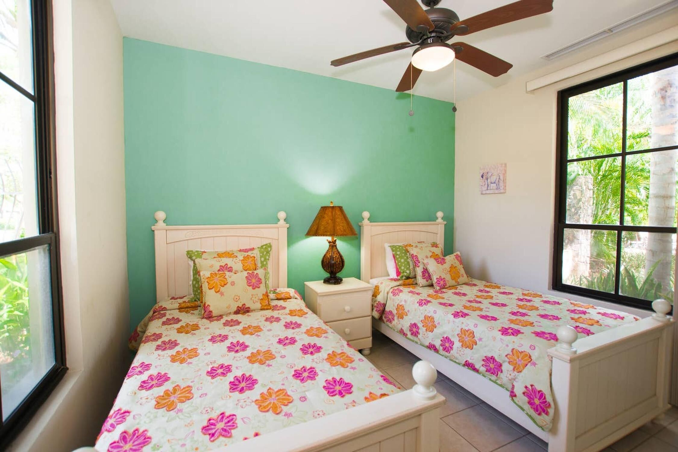 Une chambre avec deux lits simples, un ventilateur de plafond, une table de chevet avec une lampe et une fenêtre donnant sur un paysage verdoyant.