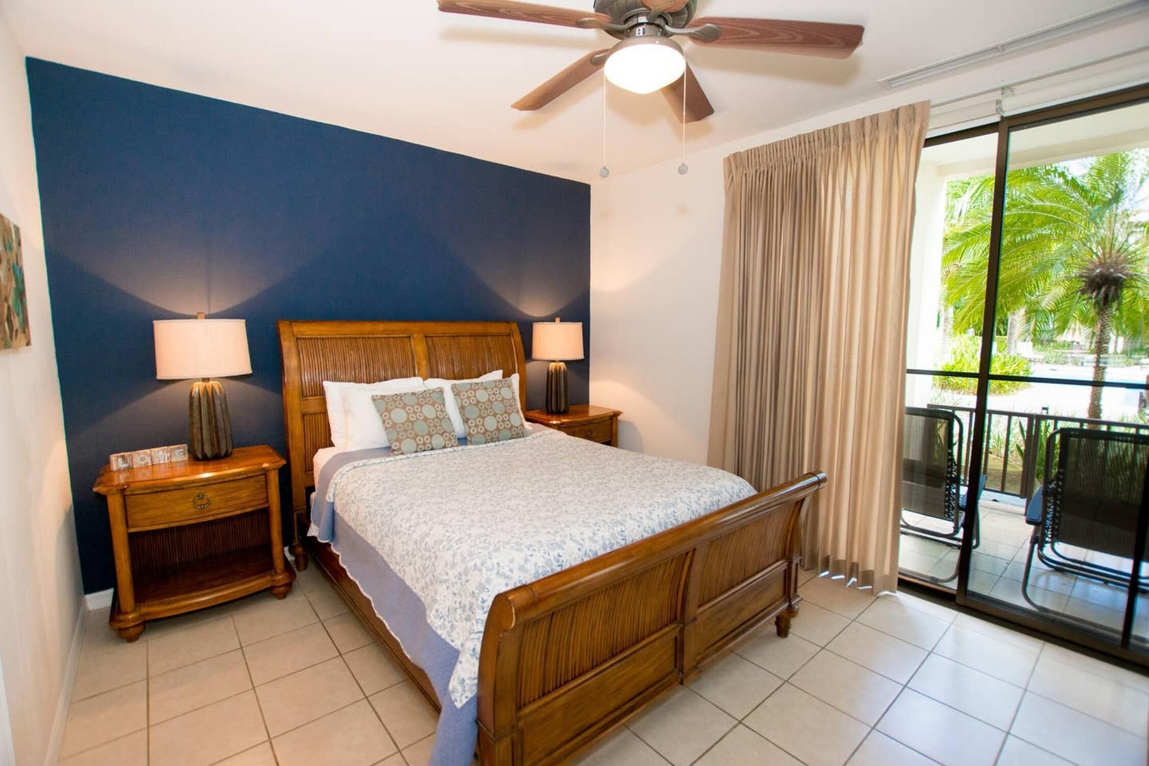 A bedroom with a wooden bed, two bedside tables with lamps, a ceiling fan, and a view of a balcony with outdoor furniture.
