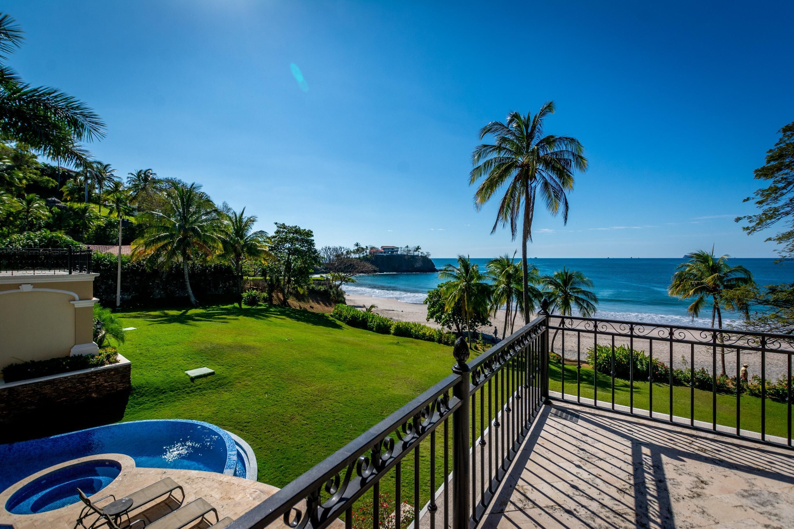 A pool with a lounge chair, a grassy lawn, and a view of the beach and ocean.