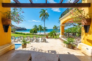 A patio with outdoor seating, a pool, and a view of the ocean and palm trees. - Elegant Mediterranean-Style Mansion with Beachfront Views and Private Infinity Pool (Cabo Velas)