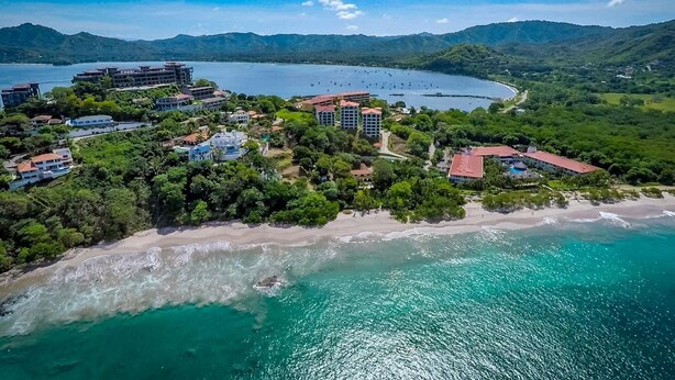 Vista aérea de um resort costeiro com vários edifícios, uma praia e uma baía.