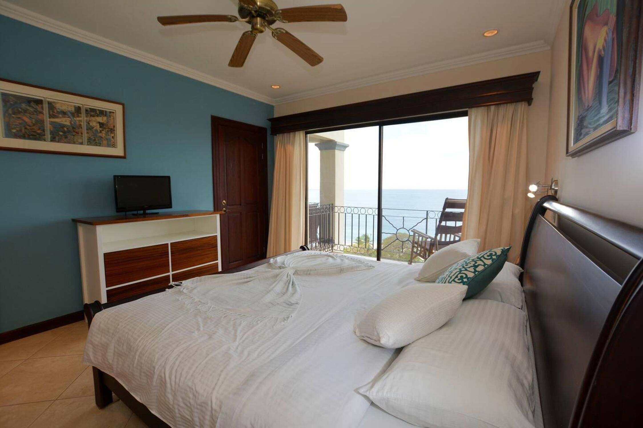 A bedroom with a bed, a television, a ceiling fan, and a balcony with a view.