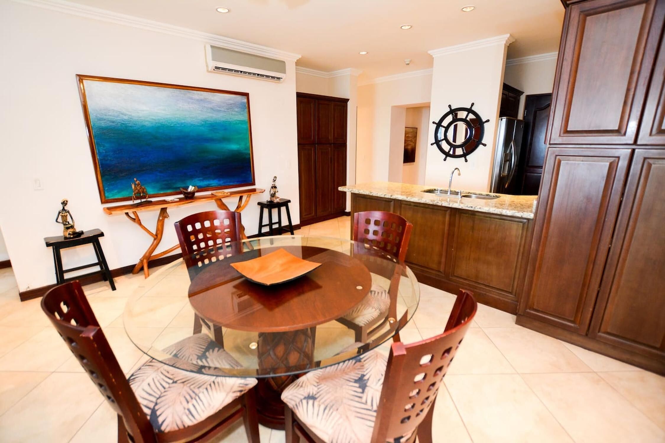 A dining area with a round glass table, wooden chairs, and a large painting on the wall.