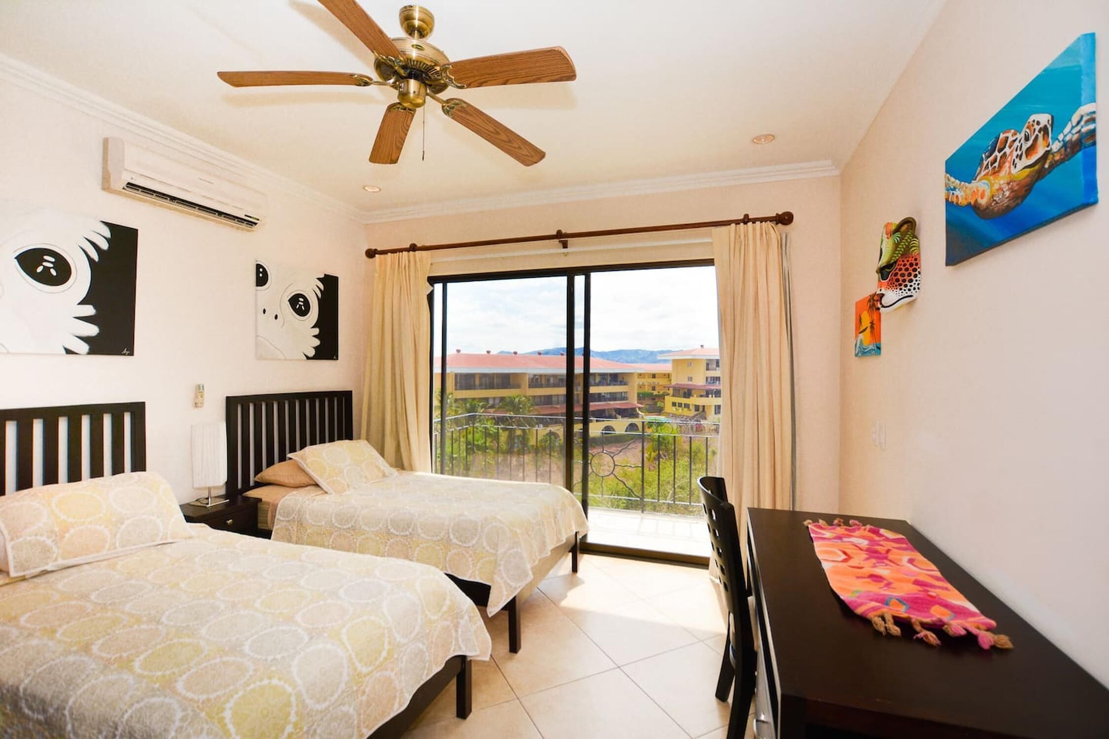 A hotel room with two beds, a ceiling fan, an air conditioner, a balcony with a view, and artwork on the walls.