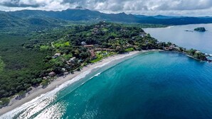 Vista aérea de una zona costera con playa, exuberante vegetación y una cadena montañosa al fondo.