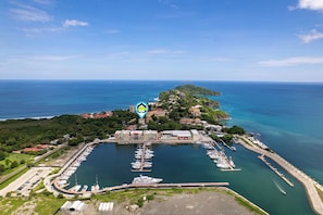 An aerial view of a marina with boats docked, a marina building, and a small island with buildings. - Top-Floor Condo w/ Sweeping Ocean, Mountain & Marina Views (Cabo Velas)