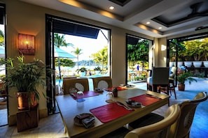 A dining area with a table set for two, large windows, and a view of an outdoor pool area. - Third-Floor Oceanview Condo with Access to Sparkling Pool, Hot Tub & Beach Club (Sardinal)