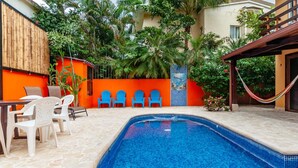 Uma piscina com azulejos azuis, rodeada por cadeiras de plástico branco e uma mesa de madeira.