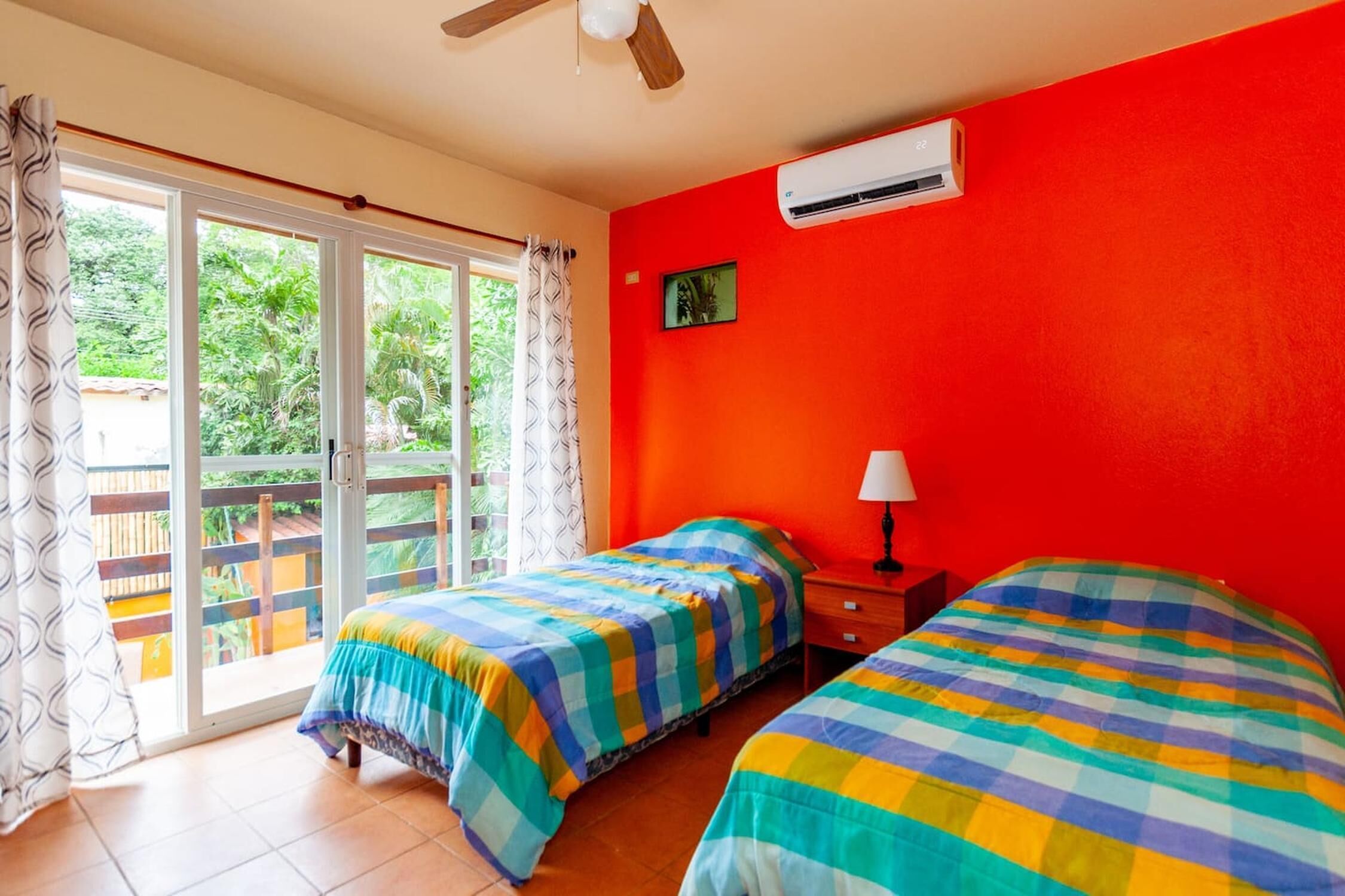 A bedroom with two beds, a red wall, an air conditioner, and a view through a glass door.
