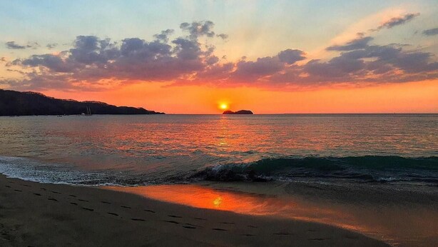 Um pôr do sol sobre uma praia com águas calmas e uma ilha ao longe.