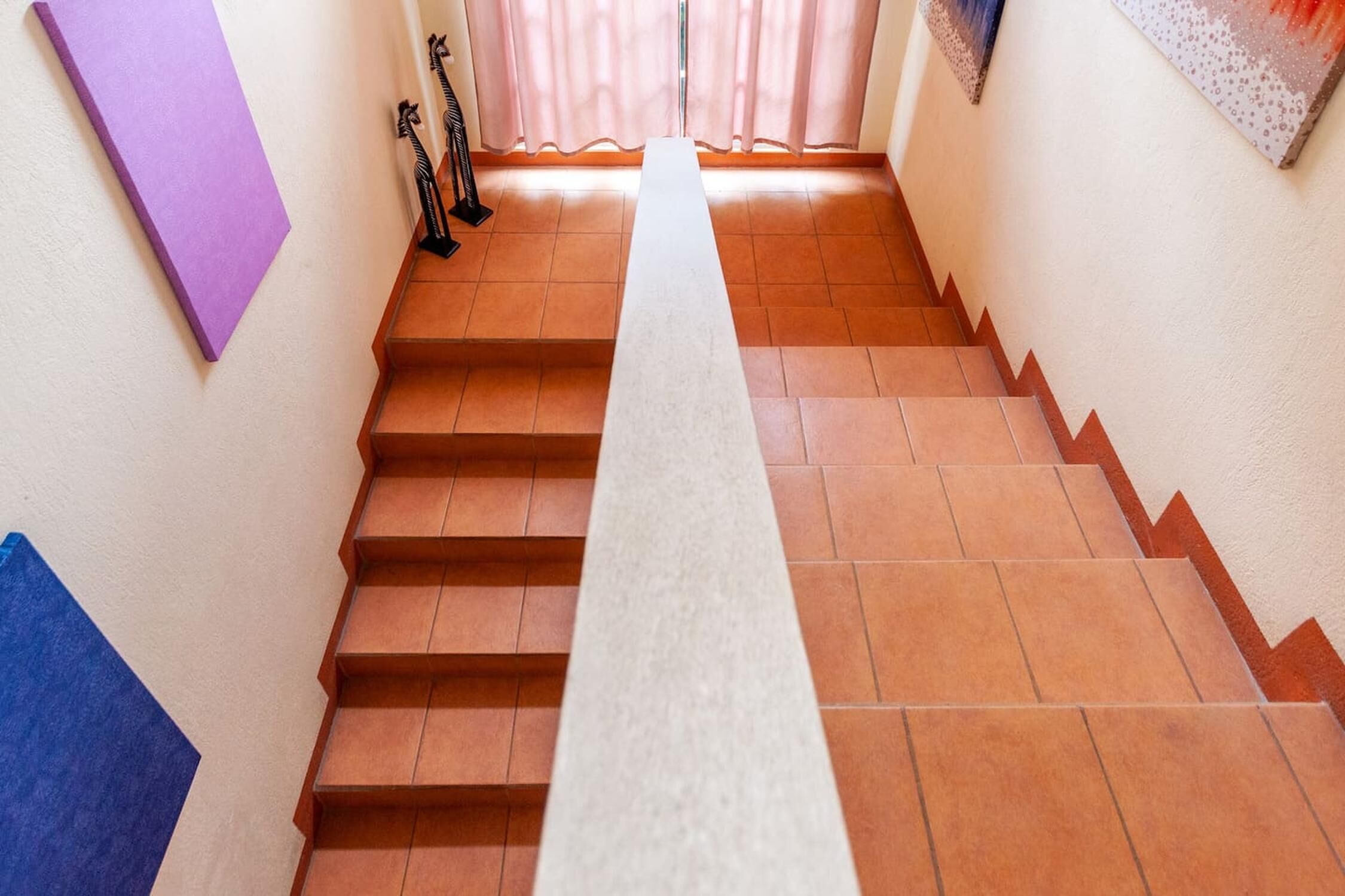 Un escalier recouvert de carreaux de terre cuite et doté d’une rampe blanche, menant à une pièce avec une fenêtre et des rideaux.
