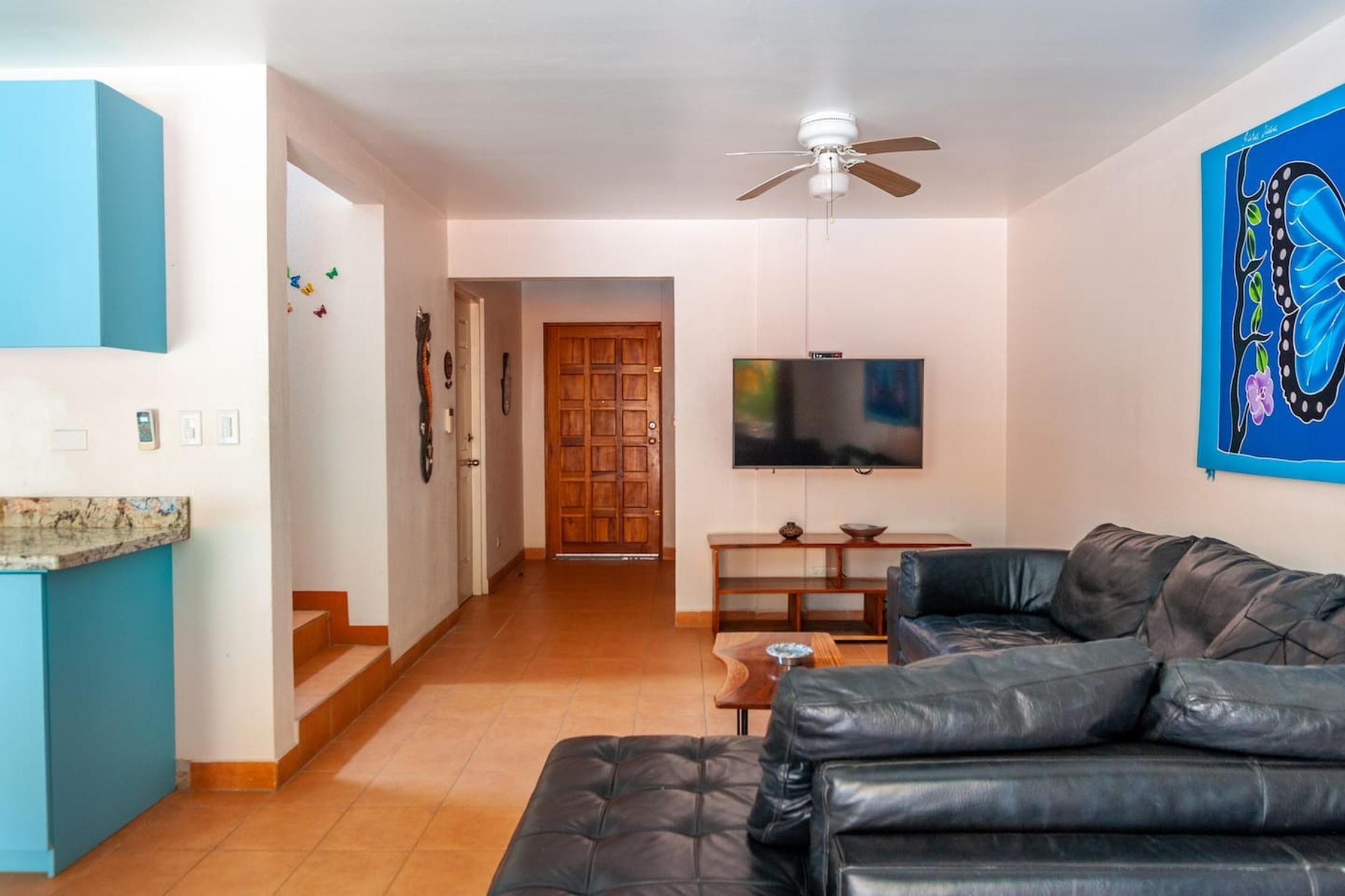 Una sala de estar con un sofá de cuero negro, un televisor montado en la pared y una puerta de madera al fondo.