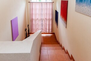 Um corredor estreito com chão em mosaico, um balcão de recepção branco, uma pasta roxa e um quadro com cores vermelhas e azuis na parede.