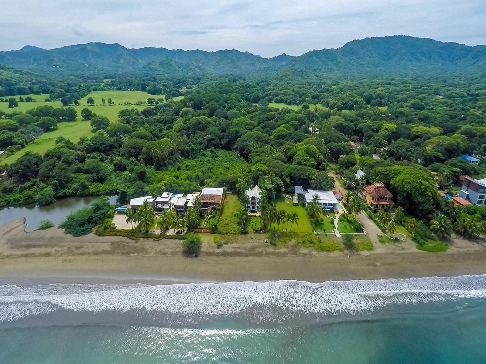 An aerial view of a coastal area with a beach, a river, and a dense forest. There are several buildings with varying roof styles, some with flat roofs and others with pitched roofs.