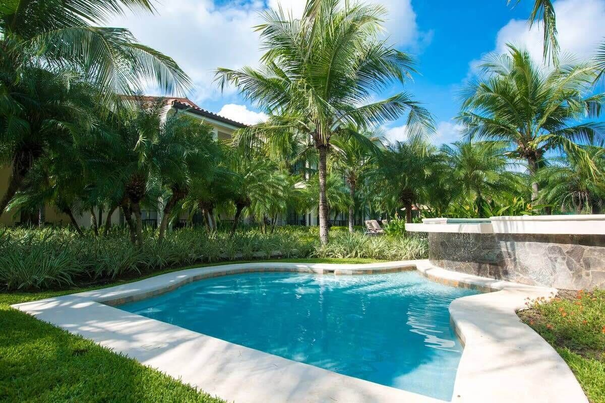 Une piscine entourée de palmiers et de verdure, avec un mur de pierre et un bâtiment en arrière-plan.
