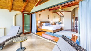 A spacious bedroom with a large bed, a sitting area with a chair and table, and a glass door leading to a balcony.