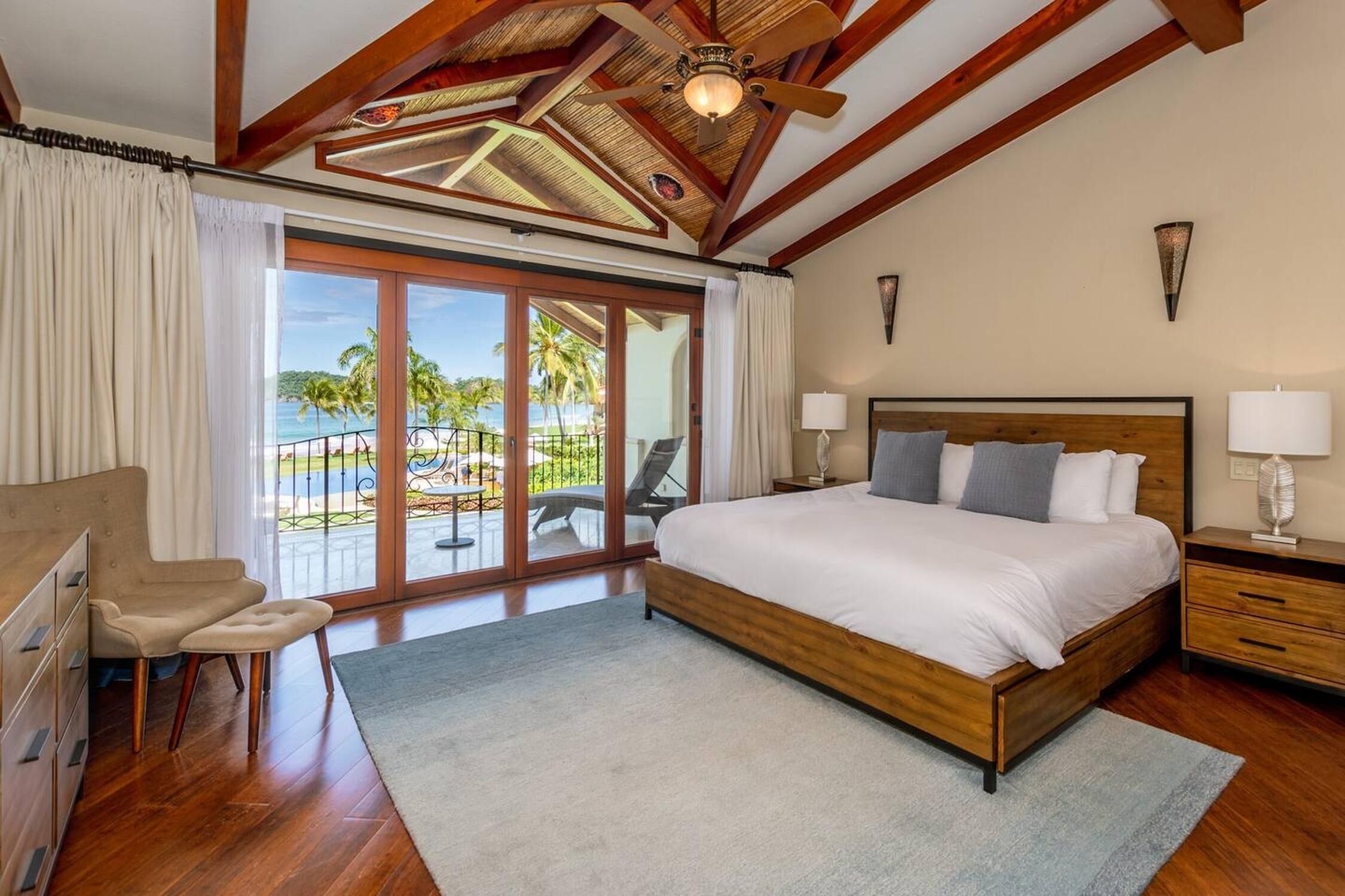 A spacious bedroom with a large bed, wooden ceiling beams, a view of the ocean, and a balcony with outdoor furniture.