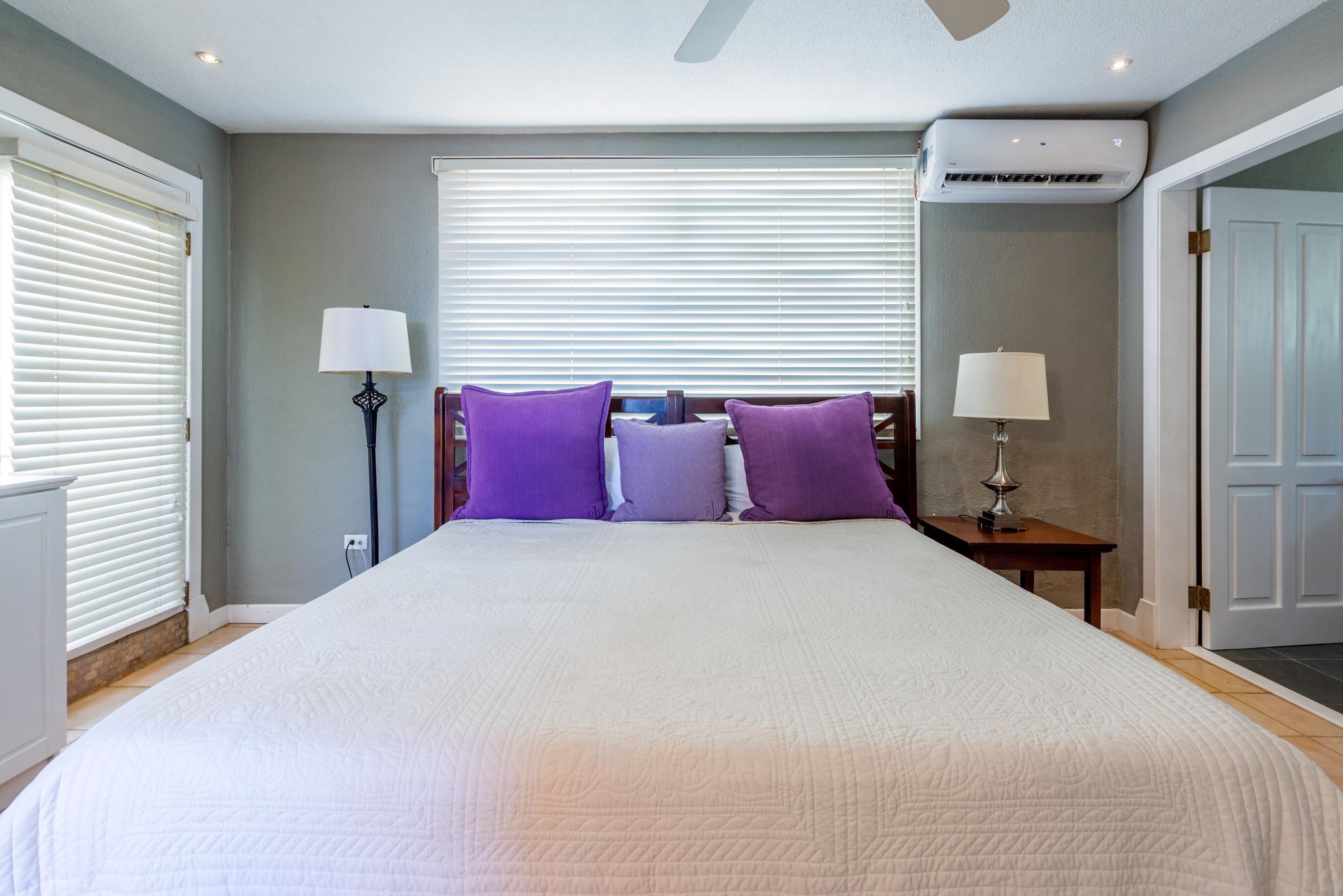 A bedroom with a large bed, two bedside tables, a ceiling fan, and an air conditioning unit.