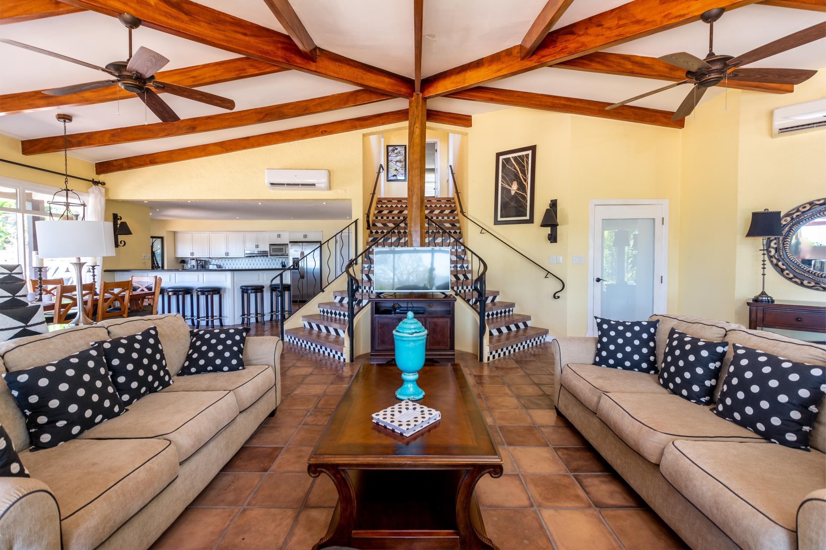 A spacious living area with a high ceiling and exposed wooden beams, a central coffee table, and a staircase leading to an upper level.