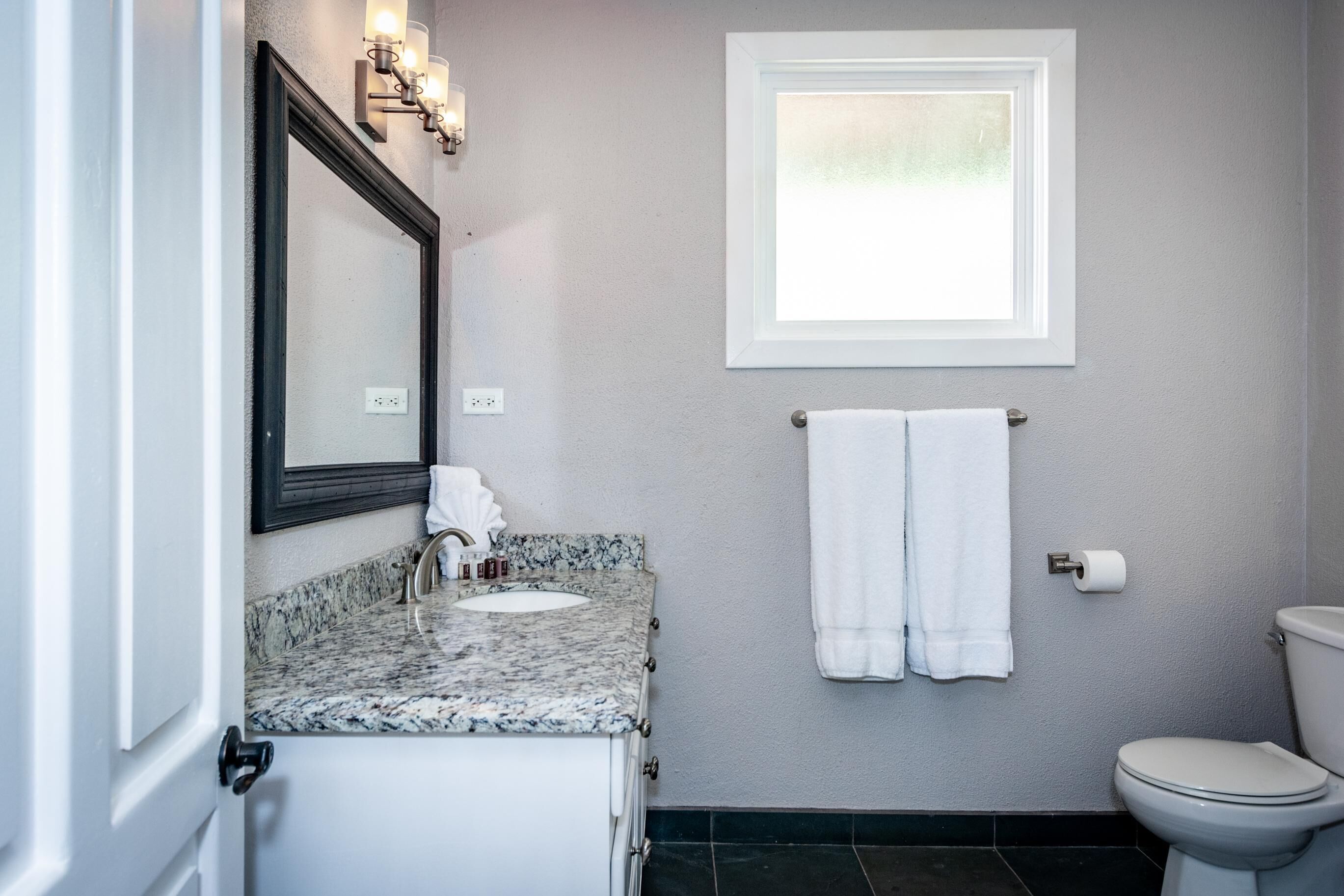 Ein Badezimmer mit Granitwaschtisch, Toilette in Weiß, einem Handtuchhalter mit zwei Handtüchern, einem gerahmten Spiegel und einem Fenster über der Toilette.