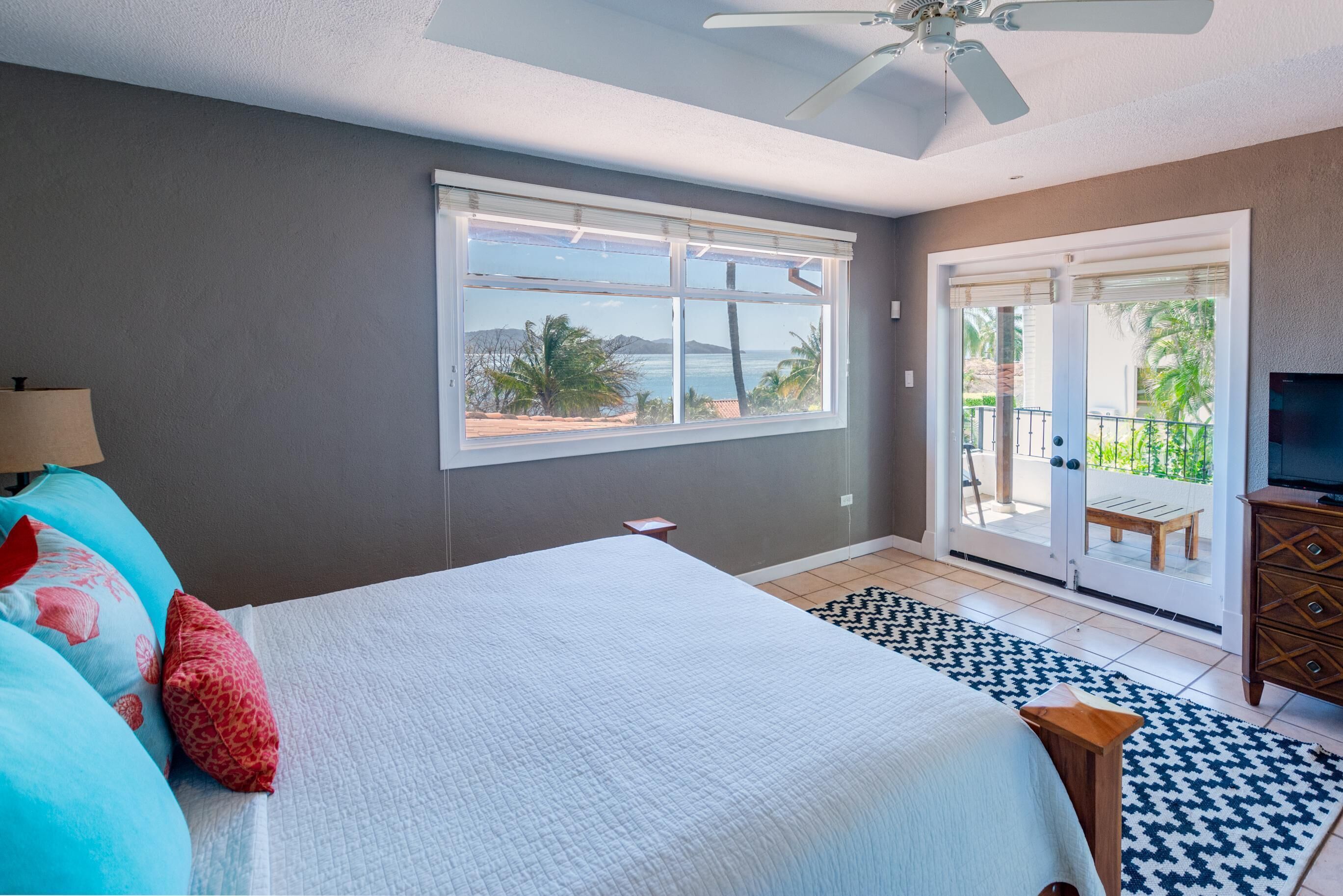 A bedroom with a bed, a TV, a ceiling fan, a window with a view, and a sliding door to a balcony.