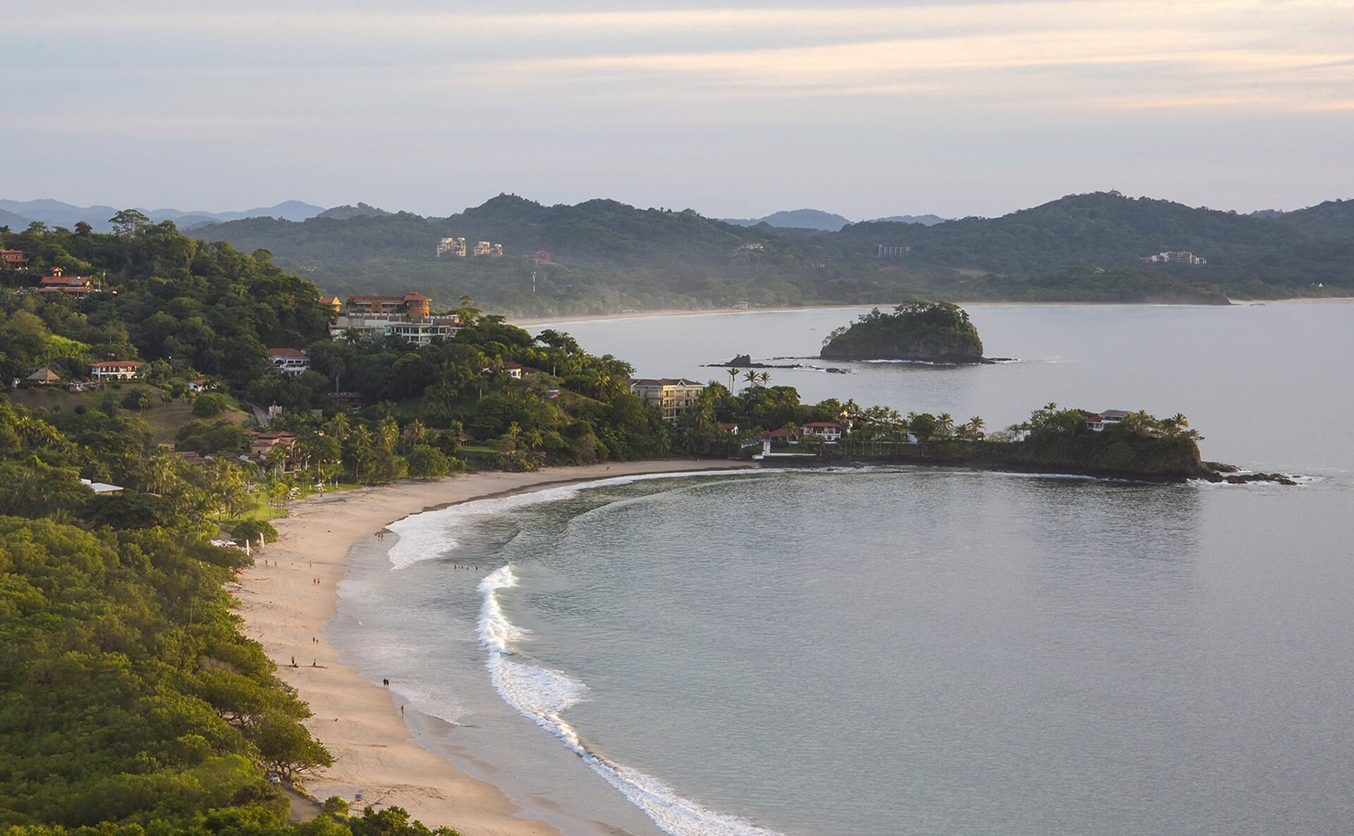 A coastal area with a sandy beach, a small island, and buildings on the hillside.