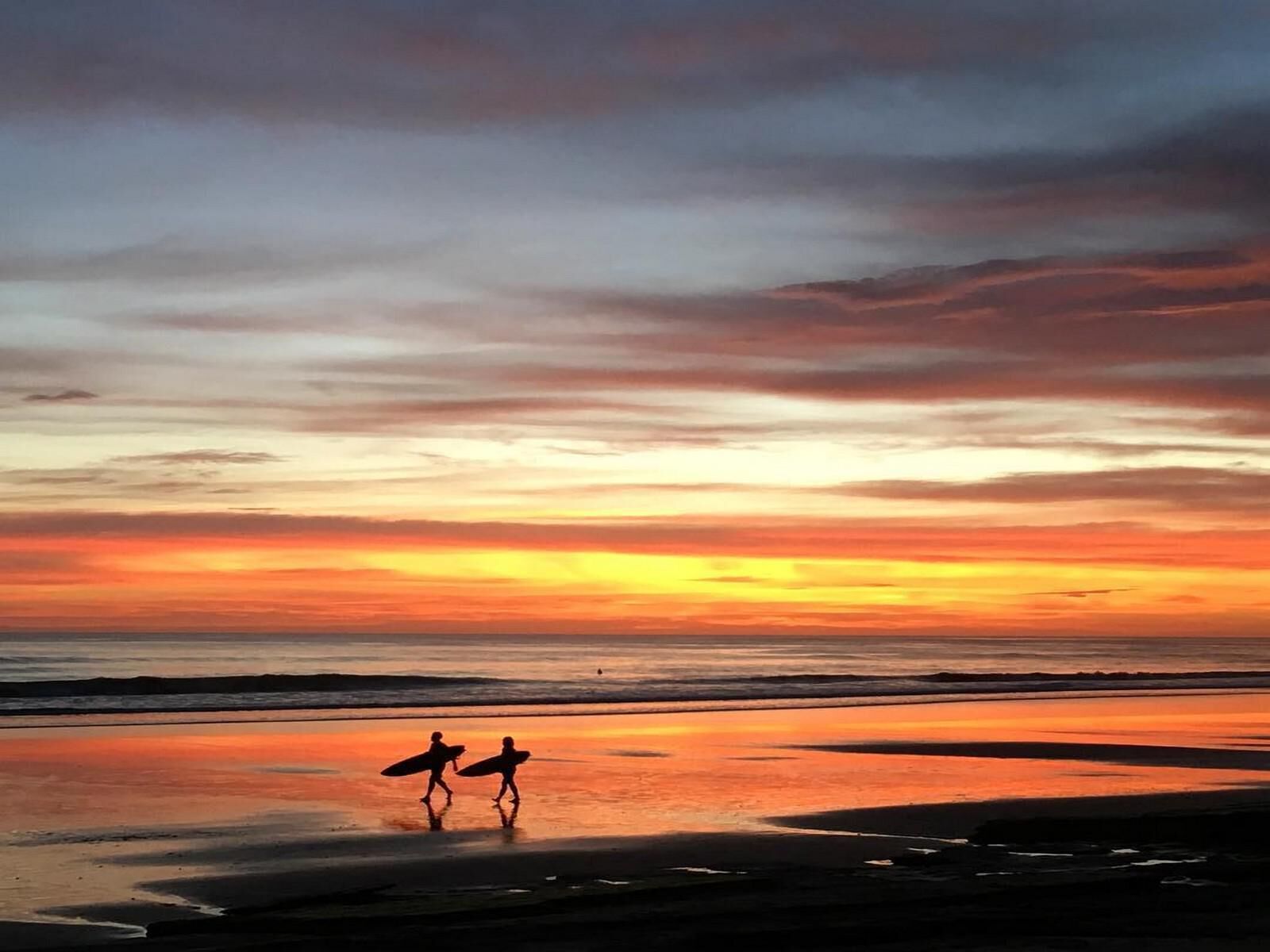 A sunset over a beach with two silhouetted figures carrying surfboards.