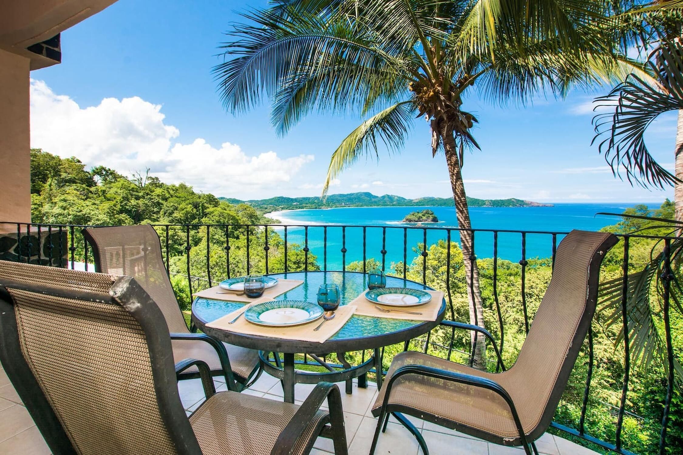Une table ronde entourée de chaises, sur un balcon donnant sur une vue panoramique de la côte.
