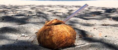 A coconut with a straw on a sandy beach with the ocean in the background.