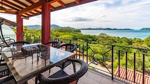 A covered outdoor area with a glass table and wicker chairs, overlooking a lush landscape and a body of water. - Stunning Hillside Villa with Panoramic Ocean Views & Infinity Pool (Tempate)