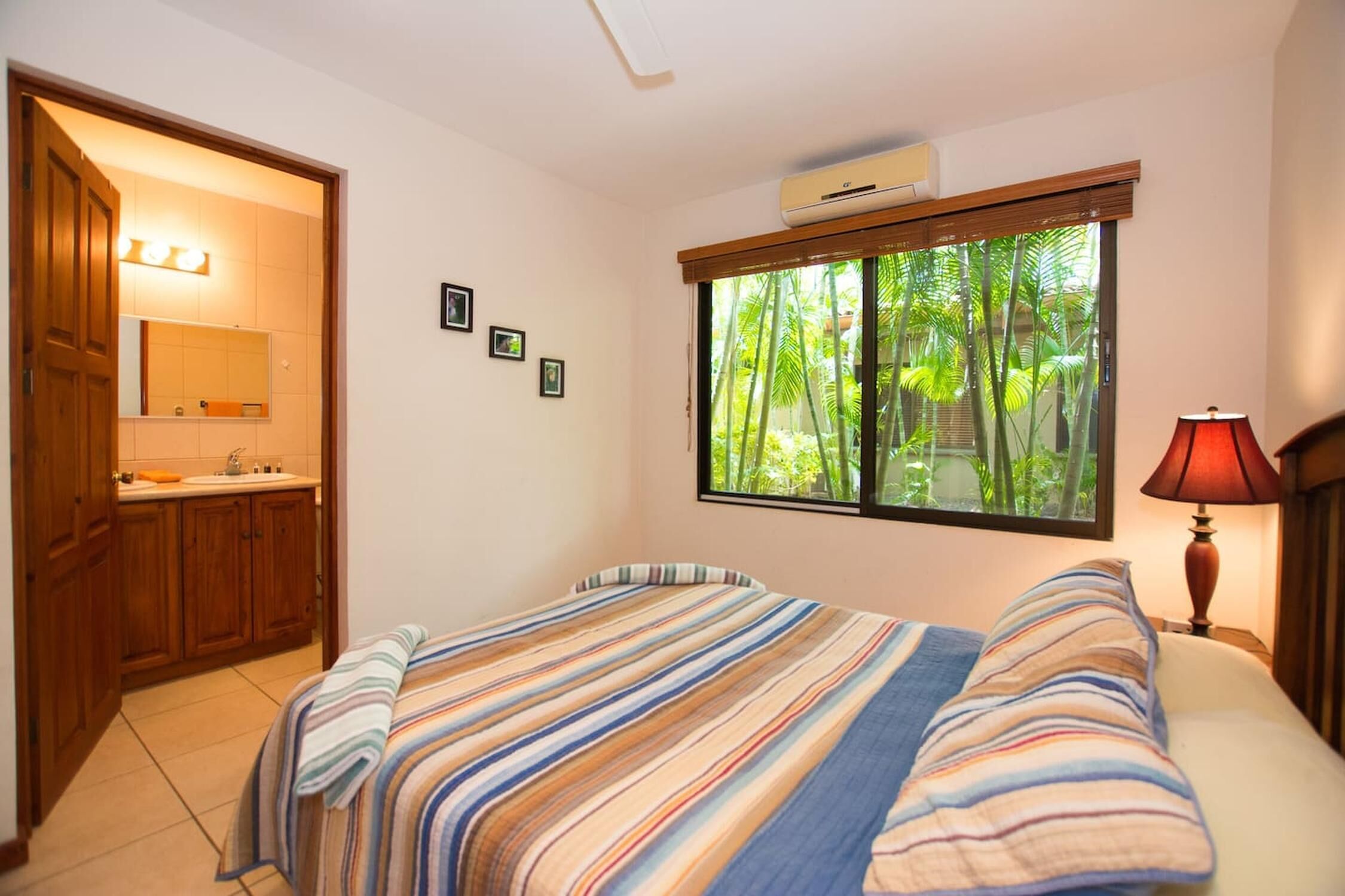 A bedroom with a bed, a window with a view of greenery, a bedside lamp, and a bathroom with a mirror and sink.